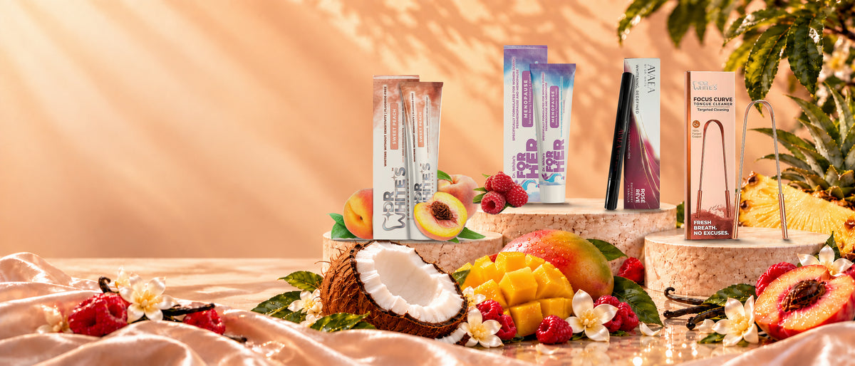 Dr. White’s Sweet Peach toothpaste, For Her toothpaste, AVAEA whitening pen and Focus Curve copper tongue cleaner on sunlit stone pedestals surrounded by oversized coconut, mango, pineapple, peach, raspberries and vanilla flowers.
